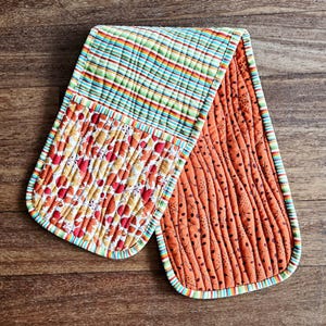 May include: A quilted oven mitt with a colorful striped top section and a patterned bottom section. The bottom section features a design of red, orange, and yellow shapes. The mitt is edged with a striped fabric.