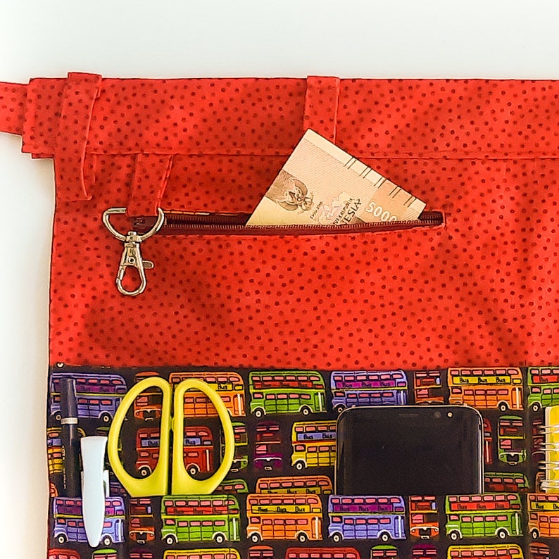 May include: Red fabric bag with a zippered pocket and a small carabiner clip. The bag has a polka dot pattern and a bottom section with a repeating pattern of double-decker buses. The bag contains a phone, a pair of scissors, a pen, and a notebook.
