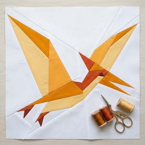 May include: A square fabric quilt block featuring a pieced geometric pterodactyl design in shades of yellow, orange, and rust on a white background, accompanied by two wooden spools of thread and gold-toned metal embroidery scissors.