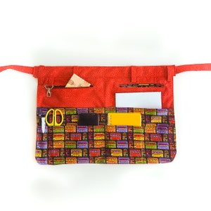 May include: Red fabric apron with multiple pockets and a colorful bus print. The apron has a zippered pocket, a small pocket for a pen, and a larger pocket for a phone or notepad.