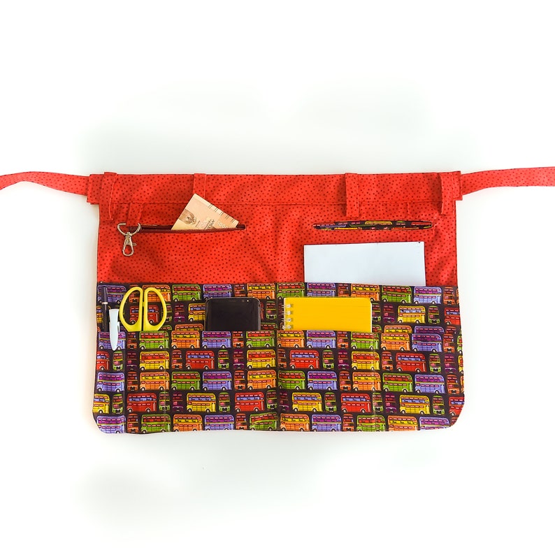 May include: Red fabric apron with multiple pockets and a colorful bus print. The apron has a zippered pocket, a small pocket for a pen, and a larger pocket for a phone or notepad.