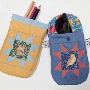 Puede incluir: Dos bolsas de tela, una amarilla y otra azul, con diseños de patchwork en forma de estrella. La bolsa amarilla contiene lápices, mientras que la azul contiene gafas de sol. Cada bolsa presenta una ilustración única y un cierre de botón.
