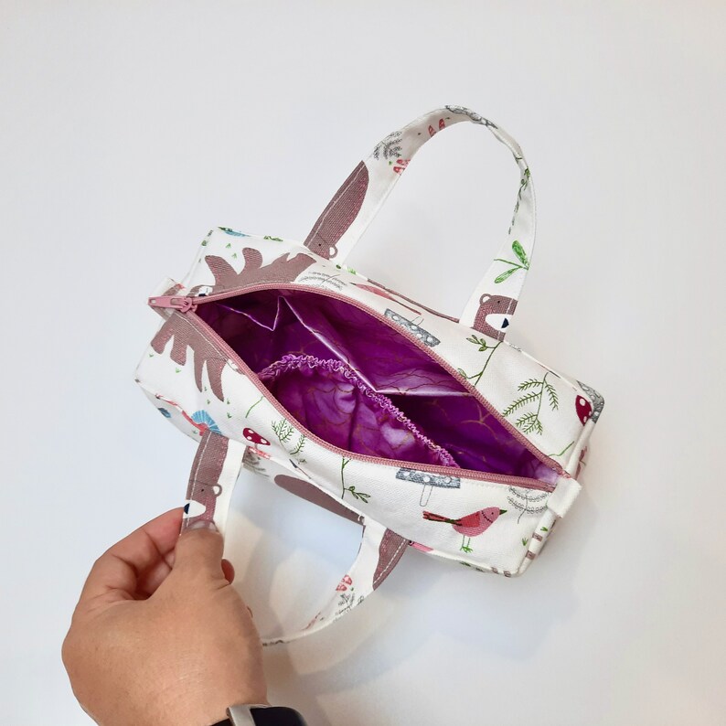 May include: A small white fabric tote bag with a pink zipper and handles. The bag is decorated with a woodland animal print featuring bears, birds, and mushrooms. The inside of the bag is lined with a purple fabric.