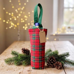 May include: A fabric wine bottle gift bag featuring a red and green plaid pattern with a solid green handle. The bag includes a blank brown paper gift tag tied with twine around the neck, holding a single bottle with a blue and silver foil top.