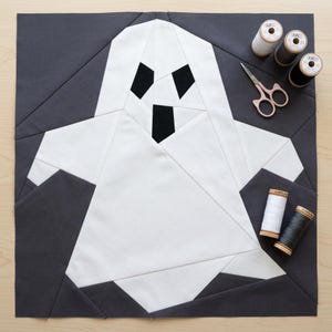 May include: A square fabric quilt block featuring a white ghost shape pieced against a dark charcoal gray background. The ghost has two black triangular eyes and a black rectangular mouth. Nearby are two white and three black thread spools and scissors.