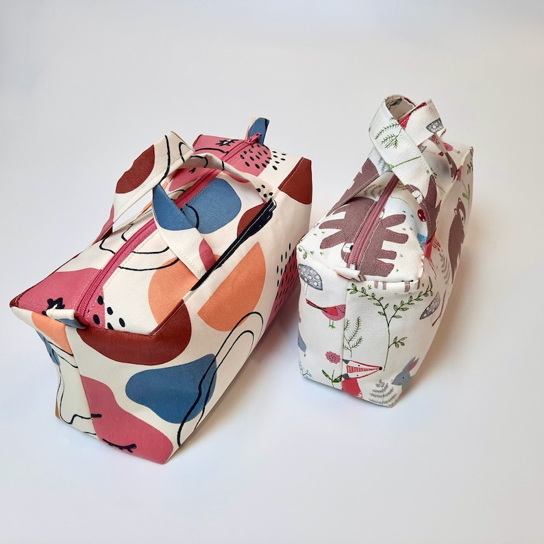 May include: Two small zippered bags with handles. One bag is white with a colorful abstract pattern in pink, orange, blue, and brown. The other bag is white with a pattern of birds, foxes, and leaves in pink, brown, and green.