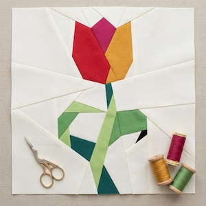 Pode incluir: Um bloco de tecido geométrico com um desenho de tulipa em vermelho, rosa e amarelo. A flor tem um caule e folhas verdes. Tesouras douradas e carretéis de linha vermelha, amarela e verde também estão presentes.