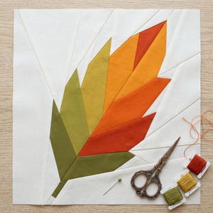 May include: A square fabric quilt block featuring a paper-pieced leaf design in shades of olive, lime, orange, and red, set against a white background. Beside the block are vintage-style metal scissors, a single green pin, and three spools of thread.
