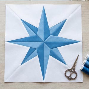 Puede incluir: Un bloque de edredón de tela cuadrado que presenta un diseño de estrella central de ocho puntas compuesto por varias piezas de tela en tonos azules sobre un fondo blanco sólido, acompañado por unas pequeñas tijeras de bordar de metal.