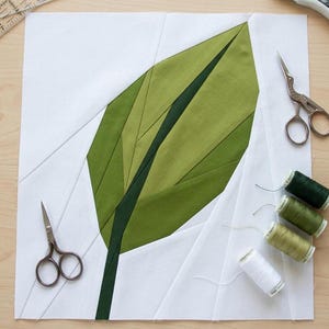 Op de afbeelding: Een stoffen blok met een groen bladerontwerp, gemaakt van verschillende tinten groen stof. Het blad staat op een witte achtergrond, met twee kleine zilveren scharen en spoelen groen en wit garen. Dit is een quiltproject.