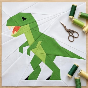 May include: A foundation paper pieced quilt block featuring a green dinosaur on a white background. The dinosaur is constructed from various shades of green fabric with a red mouth and white teeth. Bronze stork scissors and several spools of green and yellow thread are placed around the block.