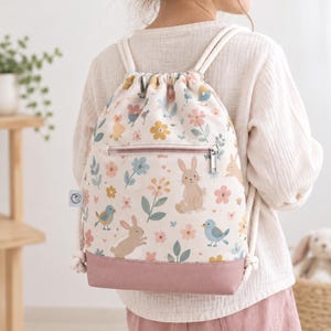 Könnte beinhalten: Ein Kordelzugrucksack aus cremefarbenem Stoff mit einem wiederkehrenden Muster aus braunen Hasen, blauen Vögeln und bunten Blumen. Die Tasche verfügt über eine horizontale Reißverschlusstasche vorne und einen festen, altrosa Boden mit dicken weißen Kordeln.
