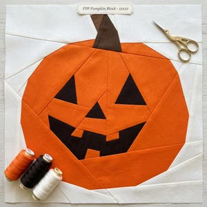 May include: A fabric foundation paper pieced pumpkin block with a white background, orange pumpkin, and black facial features. The label reads FPP Pumpkin Block - 12x12. Includes gold-toned scissors and three spools of thread in orange, black, and white.