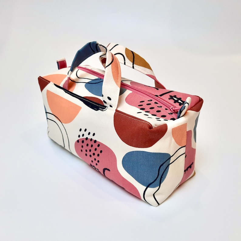 May include: A white canvas zippered tote bag with a pink, red, blue, and brown abstract pattern. The bag has two fabric handles.