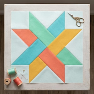 May include: A square fabric quilt block featuring a central star pattern in shades of coral, yellow, teal, and mint green, set against a pale mint background. Gold-toned embroidery scissors, two spools of thread, and a single pin are included.