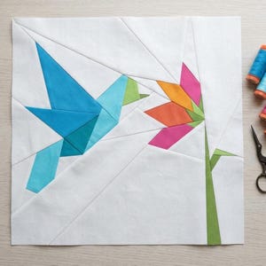 Puede incluir: Un bloque de patchwork de tela blanca con un diseño geométrico de un pájaro azul y una flor colorida. El pájaro está en vuelo y la flor tiene pétalos rosas, naranjas y verdes. El bloque está rodeado de tela blanca.