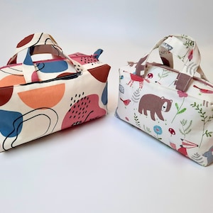 May include: Two small, rectangular zippered bags with fabric handles. One bag has a white background with abstract shapes in pink, orange, blue, and black. The other bag has a white background with a woodland pattern featuring a bear, fox, rabbit, birds, and mushrooms.
