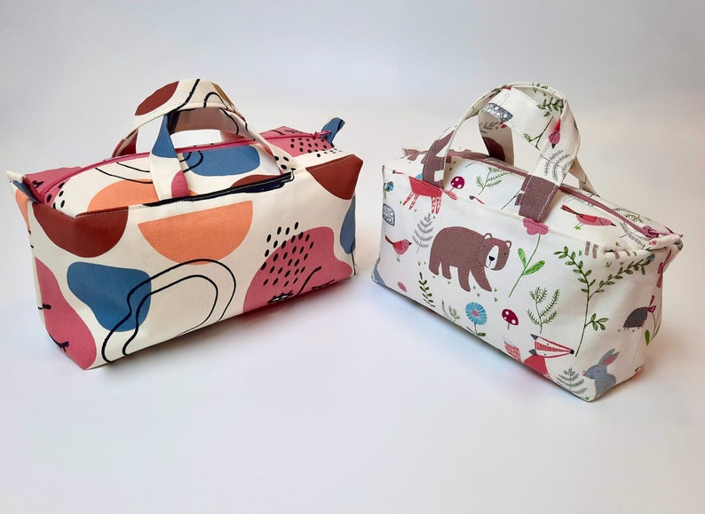 May include: Two small, rectangular zippered bags with fabric handles. One bag has a white background with abstract shapes in pink, orange, blue, and black. The other bag has a white background with a woodland pattern featuring a bear, fox, rabbit, birds, and mushrooms.