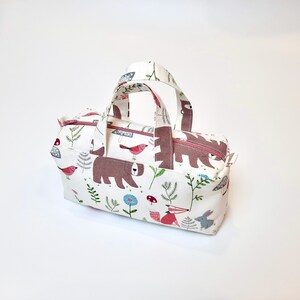May include: A small white canvas tote bag with pink handles. The bag has a woodland print featuring bears, birds, mushrooms, and flowers.