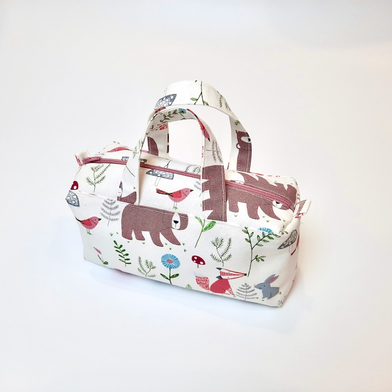May include: A small white canvas tote bag with pink handles. The bag has a woodland print featuring bears, birds, mushrooms, and flowers.