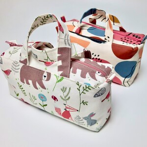 May include: Two fabric tote bags. One bag is white with a brown bear, fox, and rabbit print. The other bag is white with abstract shapes in pink, blue, and brown.