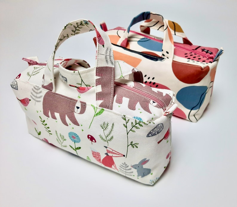 May include: Two fabric tote bags. One bag is white with a brown bear, fox, and rabbit print. The other bag is white with abstract shapes in pink, blue, and brown.