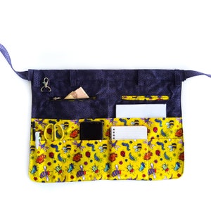 May include: A blue and yellow fabric utility apron with multiple pockets. The apron has a bee and ladybug print and includes a zippered pocket, a pen holder, a small notebook, a phone pocket, and a pocket for scissors.