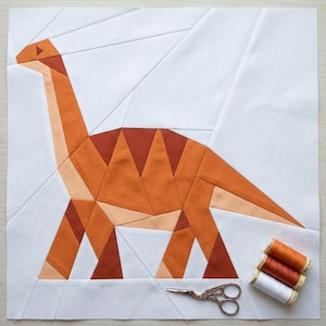 May include: A square fabric quilt block featuring a geometric paper-pieced dinosaur in shades of orange, rust, and cream on a white background. Beside the block are three spools of thread in orange, rust, and white, and metal scissors.