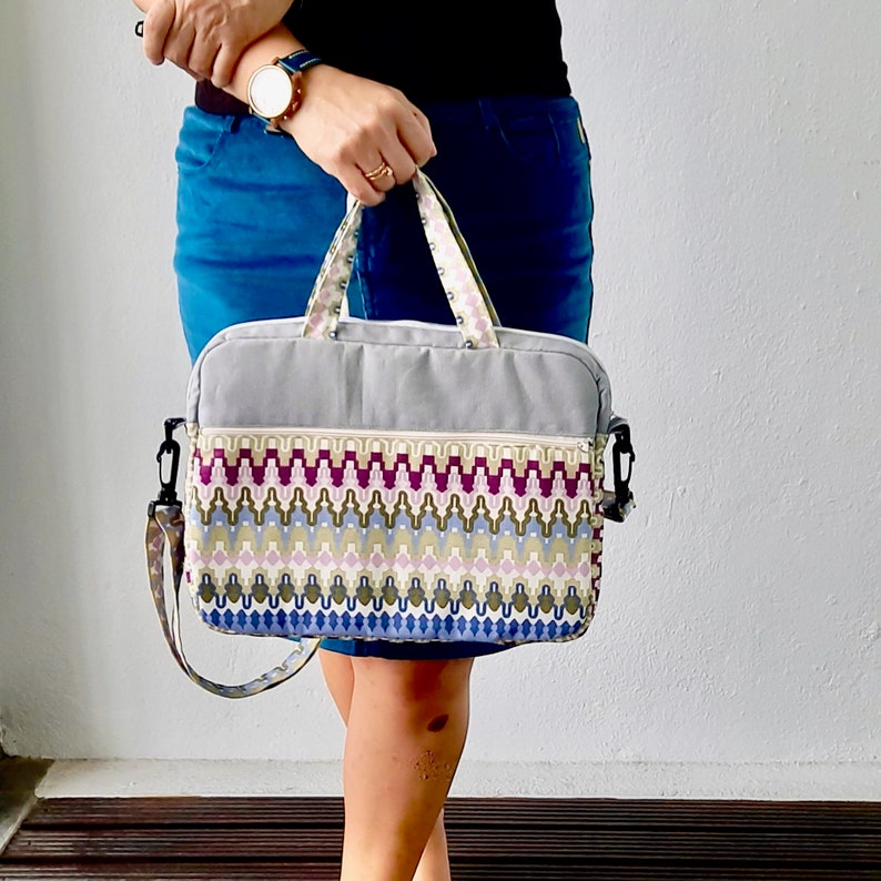K&ouml;nnte beinhalten: Eine graue Laptoptasche mit einem bunten geometrischen Muster. Die Tasche hat einen Rei&szlig;verschluss und einen oberen Griff. Die Tasche wird von einer Person getragen, die einen blauen Rock tr&auml;gt.