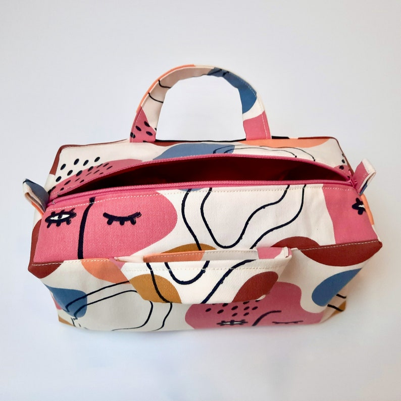 May include: A white canvas tote bag with a pink, blue, and brown abstract pattern. The bag has a zipper closure and two top handles.