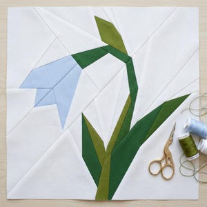 May include: A fabric block featuring a geometric design of a blue flower with green leaves and stem. The flower is set against a white background. Gold scissors and spools of thread are in the lower right corner.