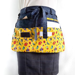 May include: A black and yellow bee-themed apron with multiple pockets. The apron has a black fabric base with a yellow fabric overlay featuring a pattern of bees, ladybugs, and caterpillars. The apron has a black ribbon tie and multiple pockets for carrying items.