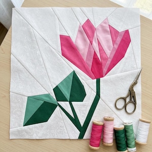 May include: A handmade fabric block featuring a geometric flower design in shades of pink and green. The flower is centered on a white background with a subtle floral pattern. A pair of scissors and spools of thread in coordinating colors are nearby.