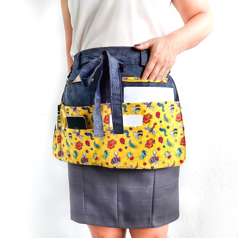 May include: A yellow and black patterned apron with multiple pockets, including a large pocket for a phone or tablet. The apron has a black waistband and a blue ribbon tie.