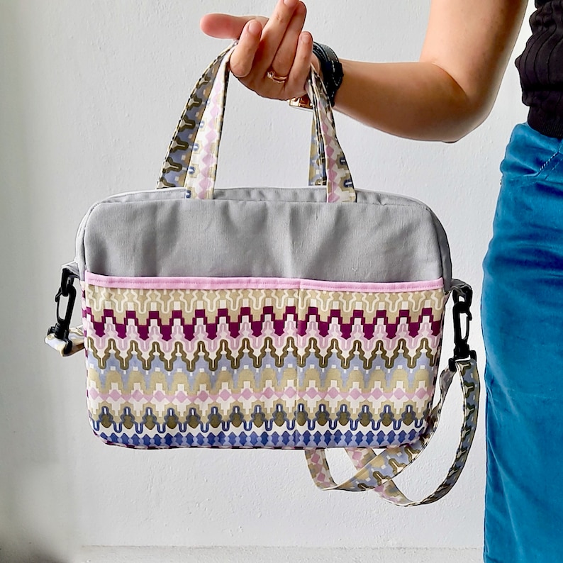K&ouml;nnte beinhalten: Eine graue Laptoptasche mit einem geometrischen Muster in Rosa und Braun. Die Tasche hat einen Tragegriff oben und einen Schultergurt.