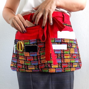 May include: Red utility apron with multiple pockets, featuring a colorful bus print. The apron has a long tie and is perfect for organizing tools and supplies.