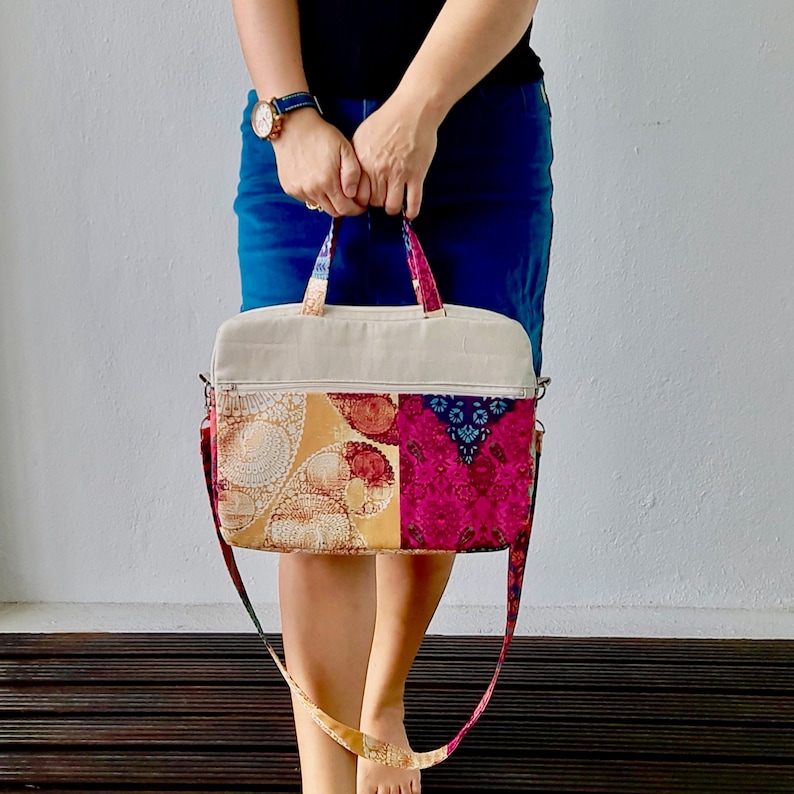 K&ouml;nnte beinhalten: Eine Patchwork-Stofftasche mit beigefarbener Canvas-Basis und einem langen verstellbaren Trageriemen. Die Tasche hat ein farbenfrohes Blumenmuster in Rosa-, Blau- und Gelbt&ouml;nen.