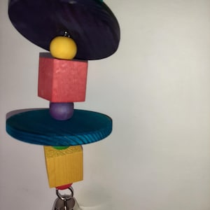 Puede incluir: Un juguete de madera colorido para pájaros con una campana. El juguete presenta piezas de madera de color morado, azul, rojo, amarillo y verde.