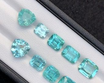 May include: A collection of nine teal green gemstones displayed in a white tray with a black border. The gemstones are various shapes and sizes, including square, rectangular, and heart shapes.