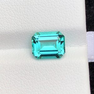 May include: A loose, emerald-cut gemstone with a vibrant, translucent cyan-green color. The stone features a rectangular shape with faceted edges that reflect light, creating a clear, brilliant appearance across its polished, multi-faceted surface.