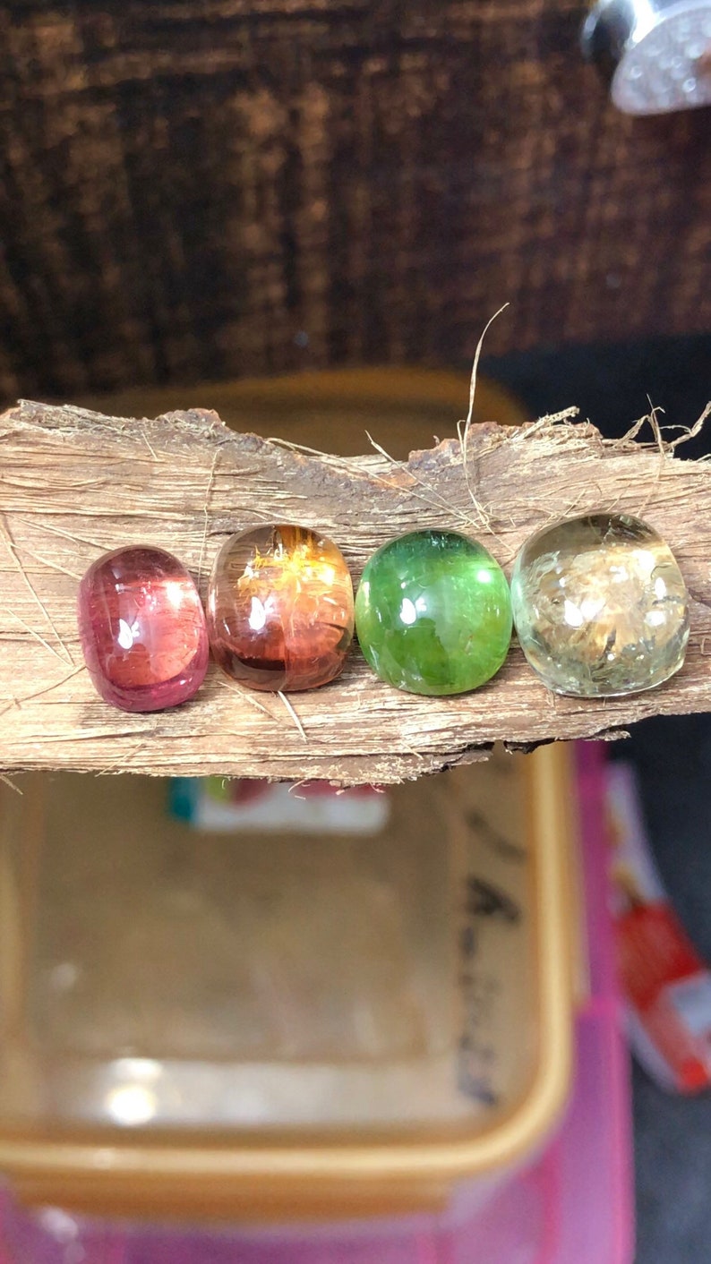 K&ouml;nnte beinhalten: Vier runde, glatte, polierte Edelsteine in verschiedenen Farben. Die Edelsteine sind in einer Reihe auf einem St&uuml;ck Holz angeordnet. Die Edelsteine sind rosa, orange, gr&uuml;n und klar.