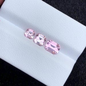 May include: Three pink gemstones, cut in a square emerald shape, are displayed on a white surface. The gemstones are arranged in a row from smallest to largest.