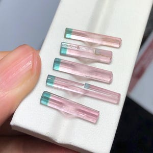 Op de afbeelding: Vijf toermalijnkristallen. Elk kristal heeft een slanke, rechthoekige vorm met een roze lichaam en een turquoise gekleurde punt. De kristallen zijn gerangschikt op een wit oppervlak, met een hand gedeeltelijk zichtbaar.