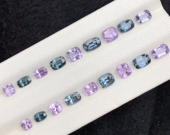 May include: A collection of 15 faceted gemstones in various shades of purple and blue. The gemstones are arranged in three rows on a white surface.