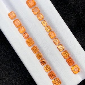 May include: A collection of twenty-one faceted orange gemstones arranged in two parallel rows on a white display stand. Each stone features a rectangular or square cut with visible internal facets that reflect light, showcasing a vibrant orange hue.