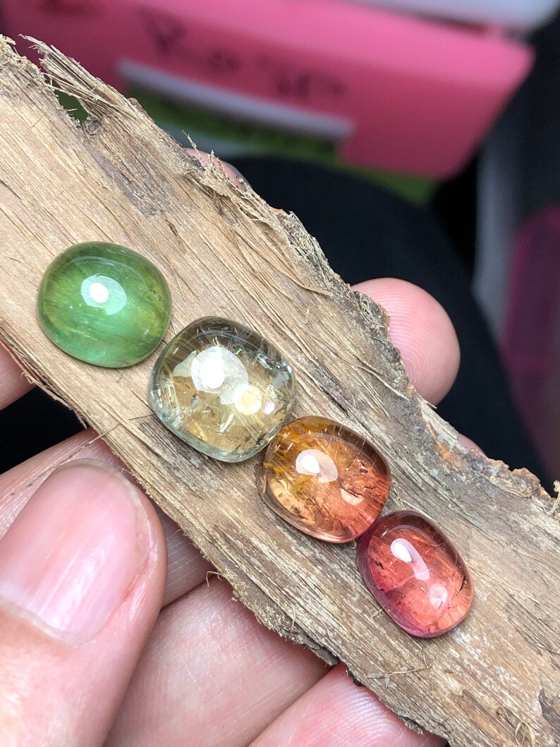 K&ouml;nnte beinhalten: Vier ovale Edelsteine in verschiedenen Schattierungen von Gr&uuml;n, Gelb, Orange und Rot. Die Steine sind in einer Reihe auf einem St&uuml;ck Holz angeordnet.