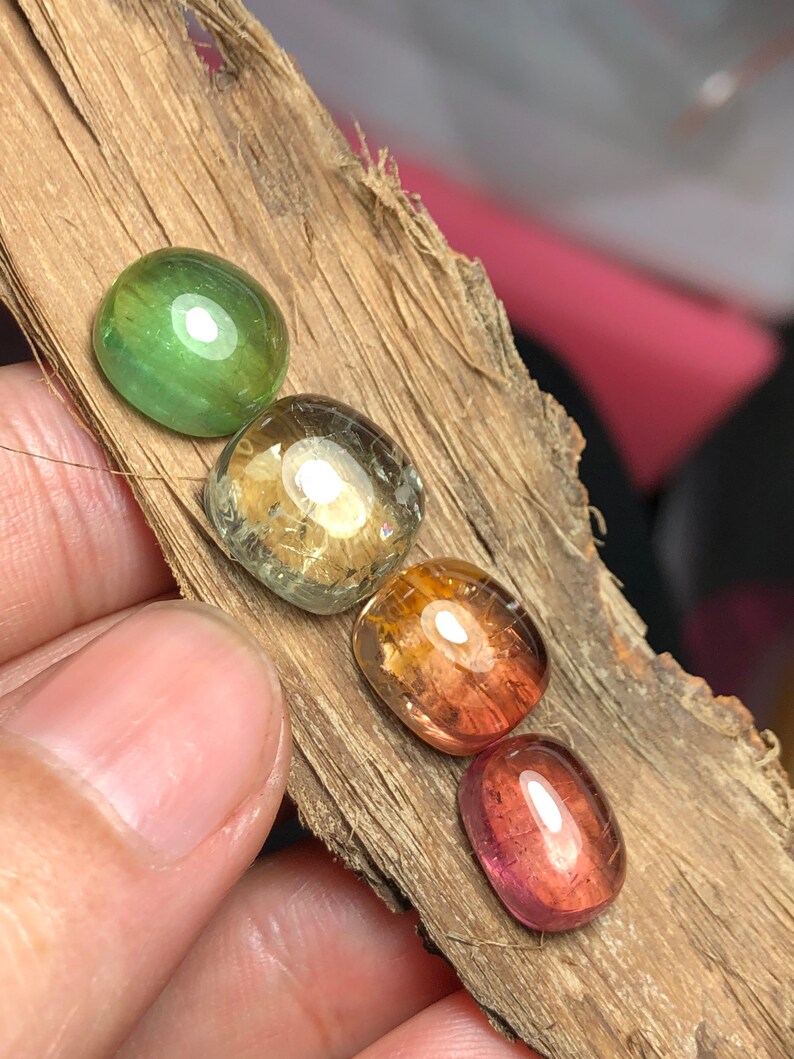 K&ouml;nnte beinhalten: Vier ovale Edelsteine in verschiedenen Farben, darunter Gr&uuml;n, Gelb, Orange und Pink. Die Steine sind in einer Reihe auf einem St&uuml;ck Holz angeordnet.