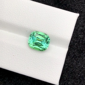 Può includere: Una gemma vibrante, taglio a cuscino, con una tonalità verde chiaro e brillante. La gemma è posta su una superficie bianca, con uno sfondo scuro. Le sfaccettature della gemma riflettono la luce, creando un effetto scintillante.