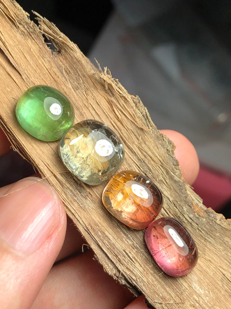 K&ouml;nnte beinhalten: Vier runde, facettierte Edelsteine in verschiedenen Farben, darunter Gr&uuml;n, Gelb, Orange und Pink, werden auf einem St&uuml;ck Holz gezeigt.