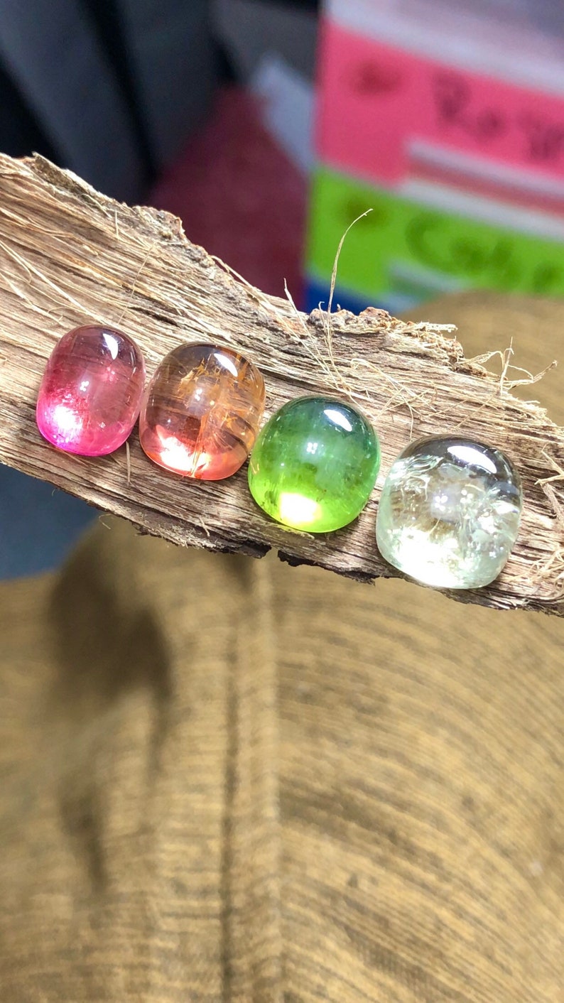 K&ouml;nnte beinhalten: Vier runde Edelsteine in verschiedenen Farben: rosa, orange, gr&uuml;n und klar. Die Edelsteine sind in einer Reihe auf einem St&uuml;ck Holz angeordnet.